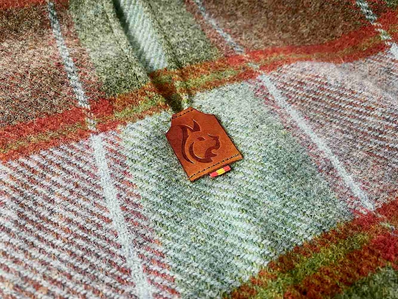 May include: A brown leather patch with a stylized animal design, attached to a plaid fabric with green, white, and orange stripes.