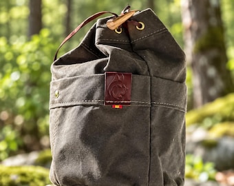 Puede incluir: Un bolso de cordón de color verde oliva oscuro hecho de tela gruesa, con un parche de cuero marrón con un logotipo de cabeza de animal en relieve, ojales metálicos de color latón y un cierre de palanca de madera en un cordón fino.