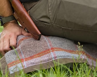 Puede incluir: Un cuchillo de hoja fija con mango oscuro está asegurado en una funda de cuero marrón lisa. La funda presenta costuras visibles a lo largo de los bordes y está colocada sobre pantalones verde oliva y una manta de lana a cuadros.