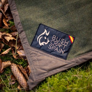 Puede incluir: Una manta de lana verde oliva con un borde de tela marrón y un parche rectangular negro con el texto BUSHCRAFT SPAIN, un logotipo de cabeza de lince y un pequeño detalle de la bandera española cosido en la esquina.
