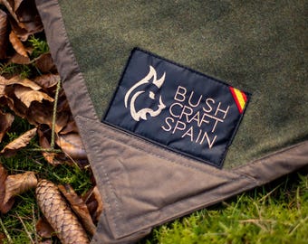 May include: An olive green wool blanket with a brown fabric border and a black rectangular patch featuring the text BUSHCRAFT SPAIN, a lynx head logo, and a small Spanish flag detail stitched onto the corner of the fabric.