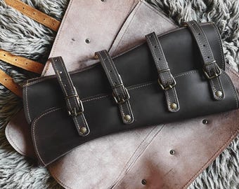 May include: A dark brown leather roll-up case featuring four adjustable straps with antique brass-toned metal buckles and rivets. The interior of the leather accessory is a lighter tan color, and the exterior is a smooth, dark brown finish.
