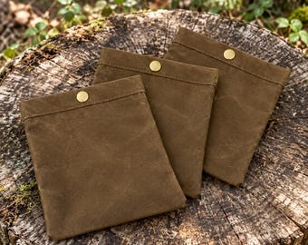 Puede incluir: Tres bolsas cuadradas hechas de tela de lona encerada en color verde oliva oscuro. Cada bolsa cuenta con un cierre de botón a presión de metal dorado centrado en el borde superior. Las bolsas están dispuestas superpuestas.