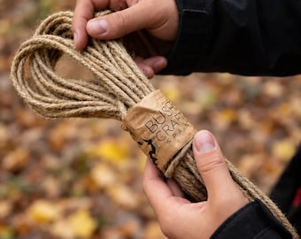 May include: A coil of natural tan jute rope secured with a brown paper band featuring the black printed text BUSHCRAFT SPAIN and a small logo. The rope is composed of multiple twisted strands and has a rough, fibrous texture throughout.