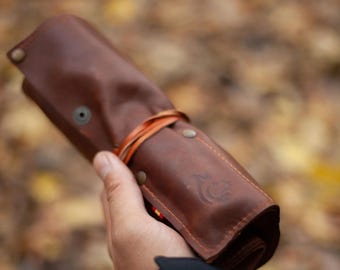 Puede incluir: Un estuche enrollable de cuero marrón asegurado con un cordón de cuero fino y botones de presión metálicos. El exterior presenta costuras visibles en los bordes y un pequeño logotipo grabado en la superficie frontal del cuero.