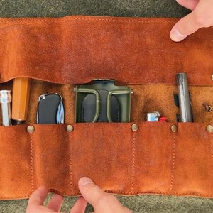 May include: A brown leather roll-up case with six slots for holding tools or other items. The case is open and shows a variety of tools, including a small knife, a pair of pliers, a screwdriver, and a small flashlight.