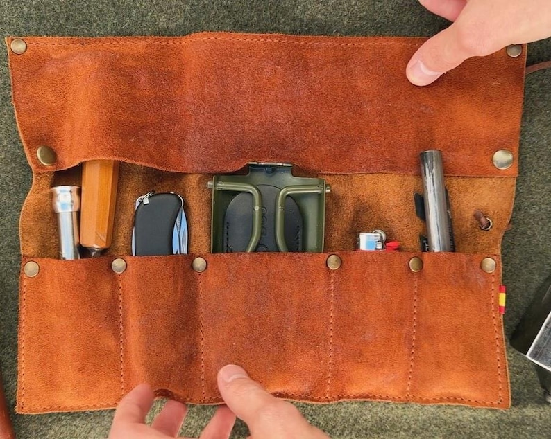 May include: A brown leather roll-up case with six slots for holding tools or other items. The case is open and shows a variety of tools, including a small knife, a pair of pliers, a screwdriver, and a small flashlight.