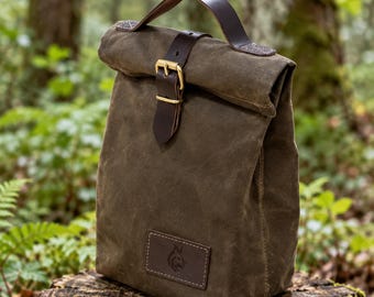 Può includere: Una borsa per il pranzo in tela cerata verde oliva con chiusura a rullo fissata da un cinturino in pelle marrone scuro e una fibbia in ottone. Un manico in pelle marrone scuro è fissato sulla parte superiore con una piccola etichetta logo.