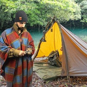 May include: A person wearing a black beanie and a plaid poncho is sharpening a knife in front of a yellow and brown tent. The tent is set up in a wooded area near a body of water.