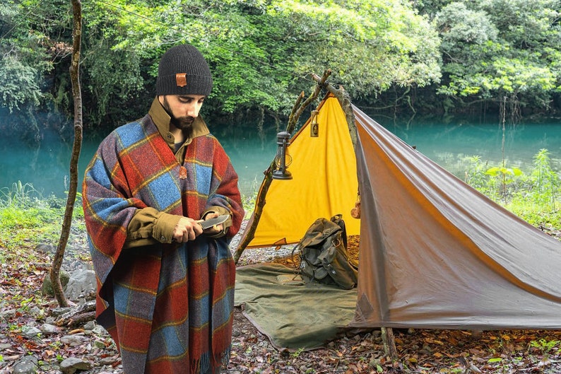 May include: A person wearing a black beanie and a plaid poncho is sharpening a knife in front of a yellow and brown tent. The tent is set up in a wooded area near a body of water.