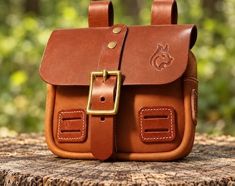 Puede incluir: Un bolso de cuero color canela con solapa frontal, cierre de hebilla de latón, dos trabillas verticales en la parte superior y dos parches rectangulares cosidos en el frente. Un pequeño logotipo de lince está grabado en la solapa.