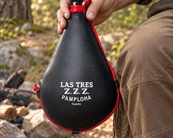 May include: A black leather wine skin canteen with a red cord trim and a black nozzle. The front features white text that reads LAS TRES Z.Z.Z. PAMPLONA España. The teardrop-shaped container is held by a black cap at the top.