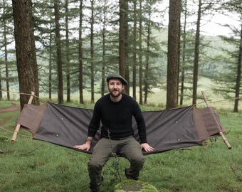 May include: A dark brown canvas hammock suspended between two wooden poles, featuring a rectangular design with metal grommets along the edges. The fabric is durable and matte, stretched horizontally to create a flat seating surface.