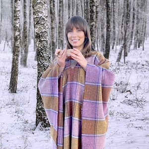 May include: A woman wearing a pink and brown plaid poncho stands in a snowy birch forest. She is holding a small wooden bowl in her hands.