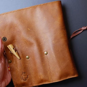 May include: A brown leather roll-up case with a flap closure and a tie. The case has three slots for holding tools or utensils. The case has a small wooden tool sticking out of one of the slots.