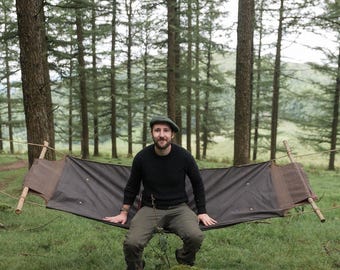 May include: A man wearing a green hat sits in a black hammock suspended between two trees in a forest. The hammock is made of canvas and has a wooden frame. The man is wearing a black sweater and green pants.