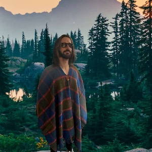 May include: A person wearing sunglasses and a blue, red, and green plaid poncho stands in front of a forest with a lake in the background.