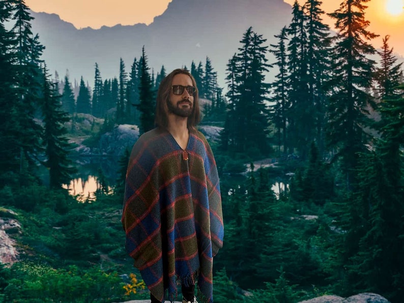 May include: A person wearing sunglasses and a blue, red, and green plaid poncho stands in front of a forest with a lake in the background.