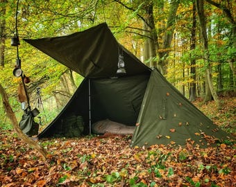 May include: An olive green canvas tarp shelter set up in a forest, featuring a metal lantern hanging from the ridge line, a sleeping bag inside, and various camping gear including a metal cup and bag suspended from a nearby tree branch.