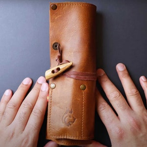 May include: A brown leather roll-up case with a wooden toggle closure and a brown leather strap. The case has a stitched design and a small animal silhouette embossed on the front.