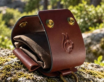 May include: A dark brown leather pouch with a curved flap secured by two brass snap buttons. The exterior features an embossed lynx head logo and a brass D-ring at the base. Inside, a soft dark brown fabric lining is visible within the folded leather structure.