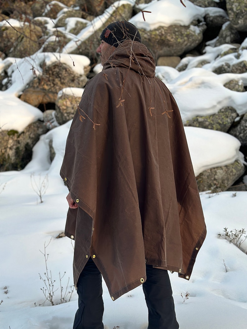 May include: A brown canvas poncho with brass snaps and a hood. The poncho is worn by a person standing in a snowy landscape.