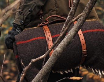 Può includere: Una coperta in lana marrone scuro arrotolata e fissata con due cinghie in pelle color cuoio dotate di fibbie in metallo e una maniglia. La coperta presenta una striscia rossa orizzontale e frange bianche alle estremità.