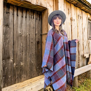 May include: A woman wearing a gray hat and a blue and red plaid poncho stands in front of a wooden cabin.