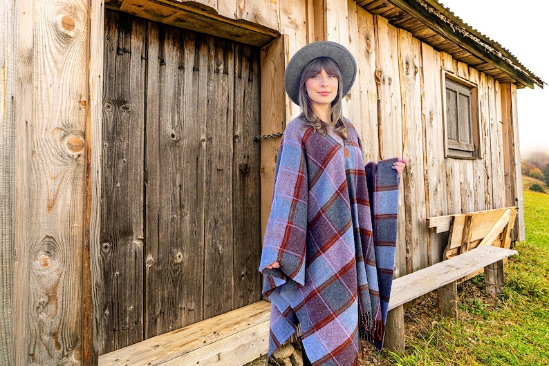 May include: A woman wearing a gray hat and a blue and red plaid poncho stands in front of a wooden cabin.