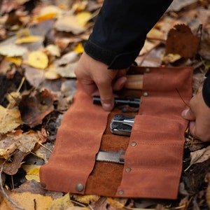 May include: A brown leather roll with multiple pockets for holding tools and knives. The roll is unrolled and shows a multi-tool, a knife, and a flashlight.