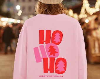 Könnte beinhalten: Ein hellrosa Langarm-Sweatshirt mit einer Grafik auf der Rückseite, die den Text HO HO HO in Rot und Pink zeigt, mit kleinen Tannenbaum-Symbolen in den Buchstaben. Der Text MERRY CHRISTMAS mit einem kleinen Herzsymbol steht darunter.