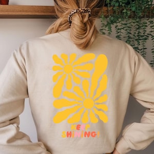 Könnte beinhalten: Beiges Sweatshirt mit einem großen gelben Blumendesign auf dem Rücken. Das Design umfasst zwei stilisierte Blumen und die Worte "KEEP SHINING" in farbigem Text. Das Sweatshirt hat eine neutrale Farbe.