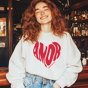 Könnte beinhalten: Ein weißes Sweatshirt mit einem großen roten Herzdesign mit dem Schriftzug "AMOR" in einer stilisierten Schriftart und "por favor" darunter. Das Sweatshirt wird mit einer blauen Jeans kombiniert. Das Bild ist in einer Bar aufgenommen.