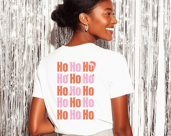 Könnte beinhalten: Ein weißes T-Shirt mit dem Text "Ho Ho Ho" in einem sich wiederholenden Muster aus Rot und Rosa. Das T-Shirt wird von einer lächelnden Person mit silbernem Hintergrund getragen.
