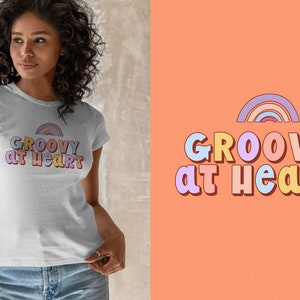 May include: White t-shirt featuring the text "GROOVY at HEART" in a colourful, retro font, with a rainbow design. The shirt is worn by a person. The background is a light orange colour.