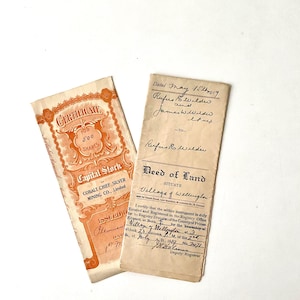 May include: Two antique documents on a white surface. One is a certificate for 500 shares from Cobalt Chief Silver Mining Co., Limited, in orange and cream. The other is a Deed of Land, with handwritten text.