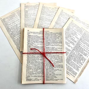 May include: A bundle of old dictionary pages, secured with red twine. Several loose pages are spread around the bundle. The pages have a yellowed, aged appearance, and the text is printed in black.