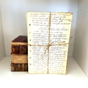 May include: A stack of handwritten notes bound with twine, featuring visible text and hole-punched edges. The notes are aged, with visible ink and a vintage aesthetic. A stack of old books is visible in the background.
