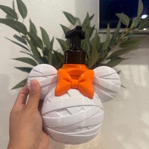 May include: A soap dispenser featuring a white mummy-wrapped character head with round ears and a bright orange bow. The dispenser has a black pump mechanism on top and an orange bottle base visible behind the white mummy-style exterior casing.