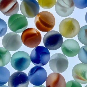 May include: A collection of colorful glass marbles in various shades of blue, green, yellow, orange, and white. Each marble has unique swirling patterns and a frosted appearance. The marbles are scattered against a bright white background.