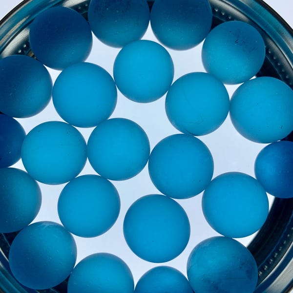 May include: A selection of blue glass marbles in various shades of blue, from light to dark, are held within a circular, teal-coloured dish. The marbles are smooth and spherical, forming an attractive arrangement.