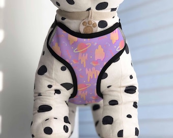 May include: A purple and orange dog harness with a space-themed pattern. The harness has a black adjustable strap and a black buckle.