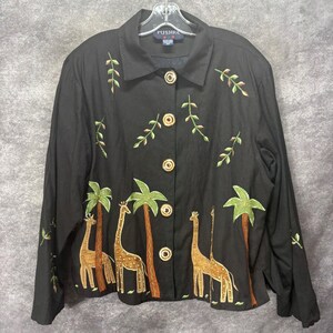 Puede incluir: Una camisa negra de manga larga con botones y un diseño bordado de jirafas y palmeras a lo largo del dobladillo inferior. La camisa tiene cuello, cinco botones redondos y pequeñas ramas de hojas bordadas en la parte superior del pecho.