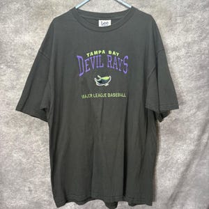 Könnte beinhalten: Ein dunkelgraues Kurzarm-T-Shirt mit dem Text TAMPA BAY DEVIL RAYS in Lila und Limettengrün, einer Rochen-Grafik und den darunter gedruckten Worten MAJOR LEAGUE BASEBALL. Das T-Shirt hat ein Lee-Markenetikett am inneren Halsausschnitt.