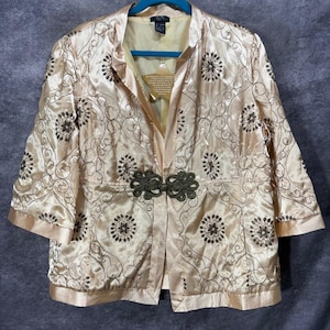 Könnte beinhalten: Eine champagnerfarbene Satinjacke mit einem komplizierten dunkelbraunen gestickten Blumenmuster und geschwungenen Linien. Das Kleidungsstück hat Dreiviertelärmel und einen einzelnen dunkelbraunen dekorativen Knebelverschluss vorne.
