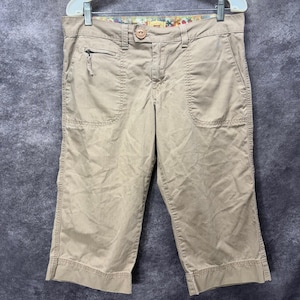 May include: Beige capri pants featuring a button and zipper fly closure, belt loops, and two front pockets. One pocket includes a small zippered compartment. The interior waistband is lined with a colorful floral patterned fabric.