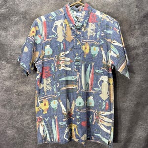 Puede incluir: Una camisa de manga corta con botones en un tono azul medio que presenta un estampado integral de varias criaturas marinas, incluyendo calamares, peces y formas marinas abstractas en tonos blanco, amarillo, rojo y azul claro con cuello en punta.