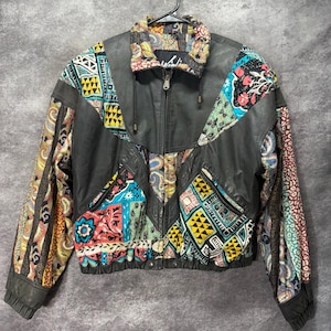 May include: A black leather jacket featuring a full-length front zipper, a high collar, and long sleeves. The garment is accented with multi-colored fabric panels displaying various geometric, floral, and paisley patterns in shades of yellow, blue, red, and teal.
