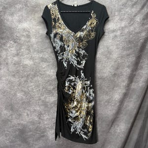 May include: Black V-neck dress with cap sleeves and a side slit. The dress features an ornate design in gold and silver tones across the front. The dress is hanging on a black hanger.