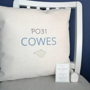 May include: Cream cushion featuring the text "PO31 COWES" in blue, with a grey island outline. The cushion rests on a white chair with a grey seat. A small white card and a white house-shaped ornament are also present.